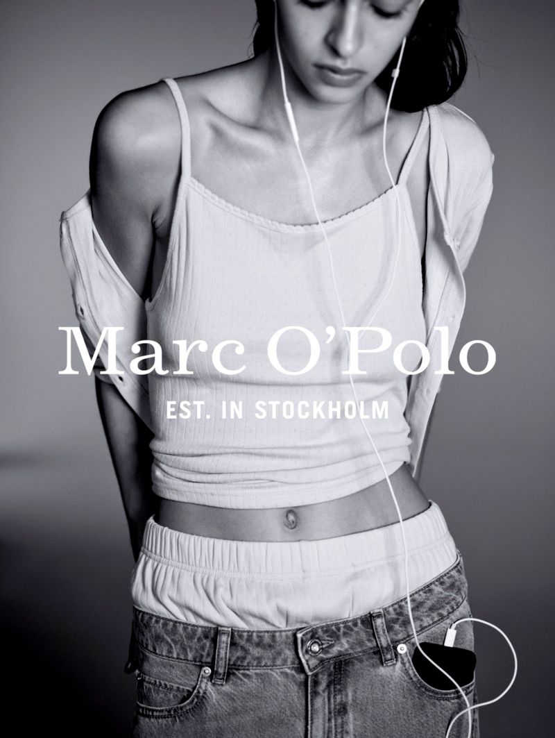 A woman poses in a casual outfit with a top, jeans, and headphones. The image features the Marc O'Polo logo, highlighting a fashion editorial aesthetic.