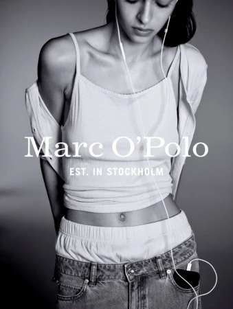 A woman poses in a casual outfit with a top, jeans, and headphones. The image features the Marc O'Polo logo, highlighting a fashion editorial aesthetic.