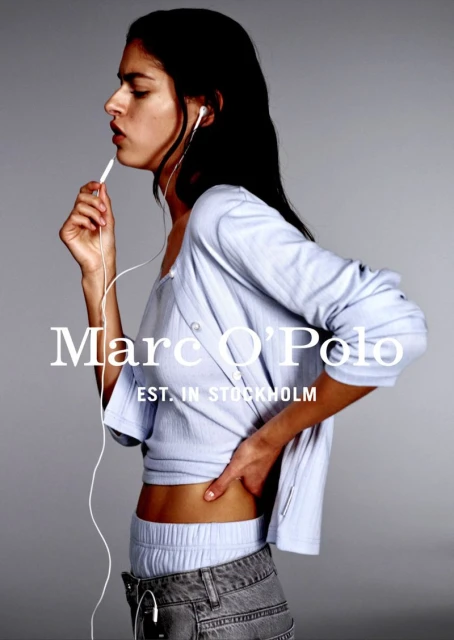 Fashion shot of a young woman wearing a blue top and jeans, listening to white earphones against a grey backdrop, with visible brand logo.