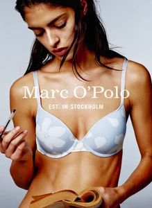 Model wearing a blue patterned bra holding a pen and book against a grey seamless background for Marc O'Polo advertising.