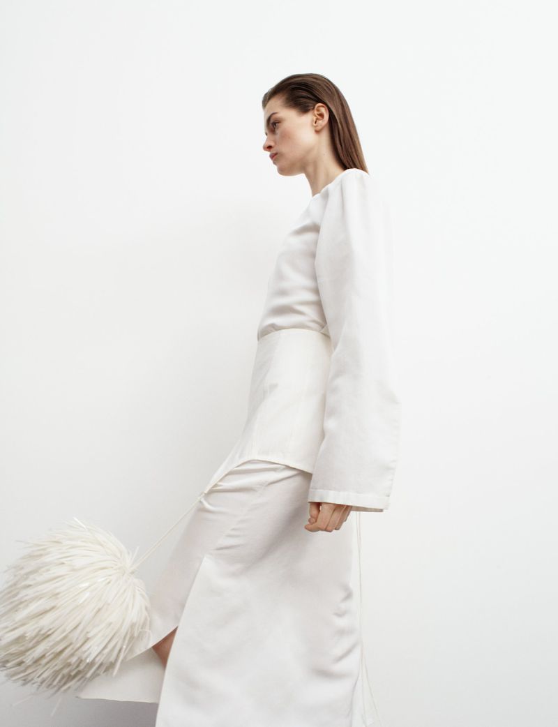 Profile of a woman wearing a white long-sleeve top and long skirt with a white furry clutch, posing in front of a white background.