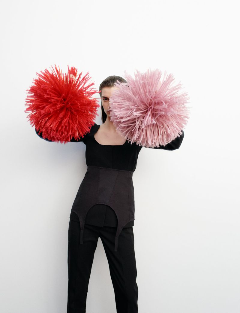 Woman in black outfit poses holding red and pink feather pom-poms against a white backdrop in a fashion editorial-style image.