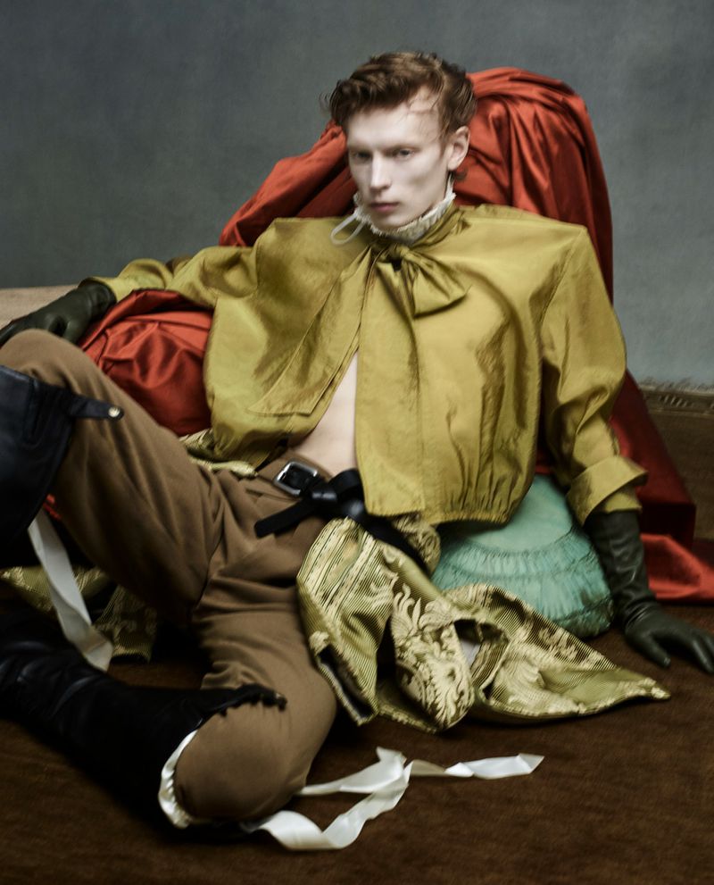 Young man poses indoors wearing a golden silk top, brown pants, black gloves and boots while surrounded by silk pillows and fabric.