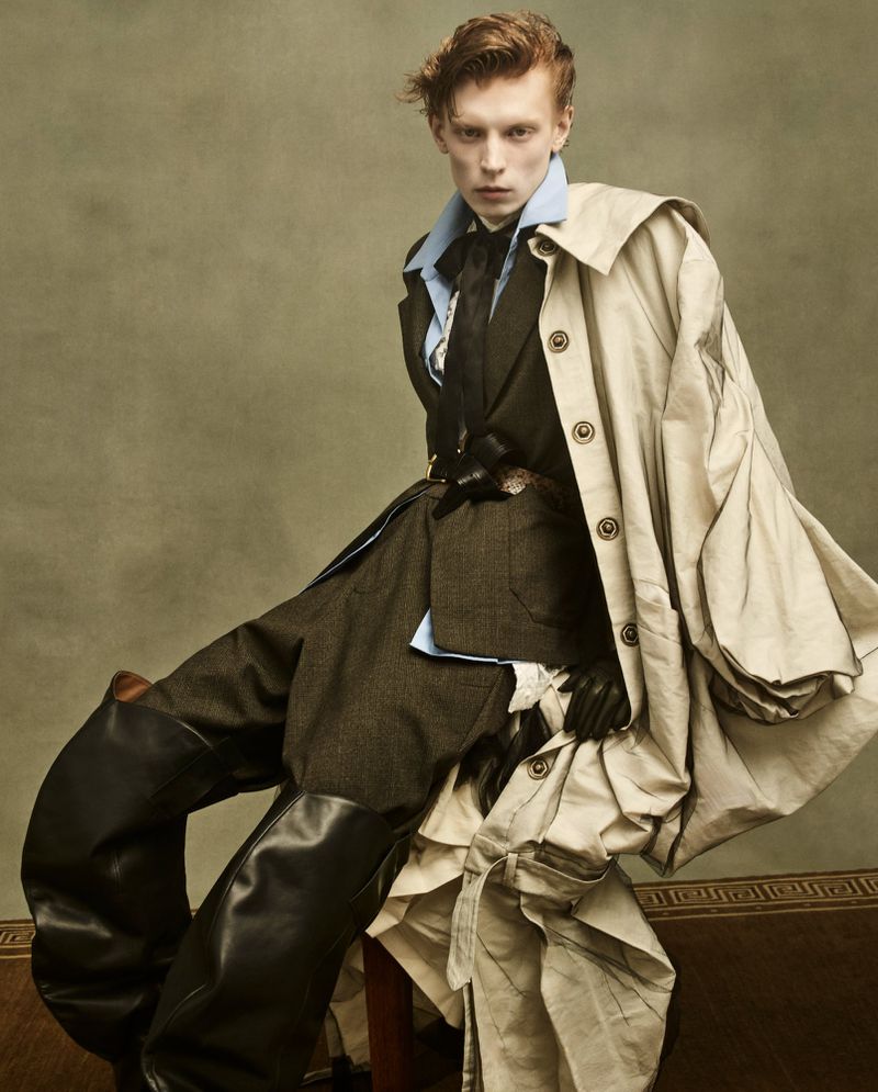 A fair-skinned young male fashion model poses indoors wearing an olive suit, leather boots, an off-white trench coat, and a black tie.