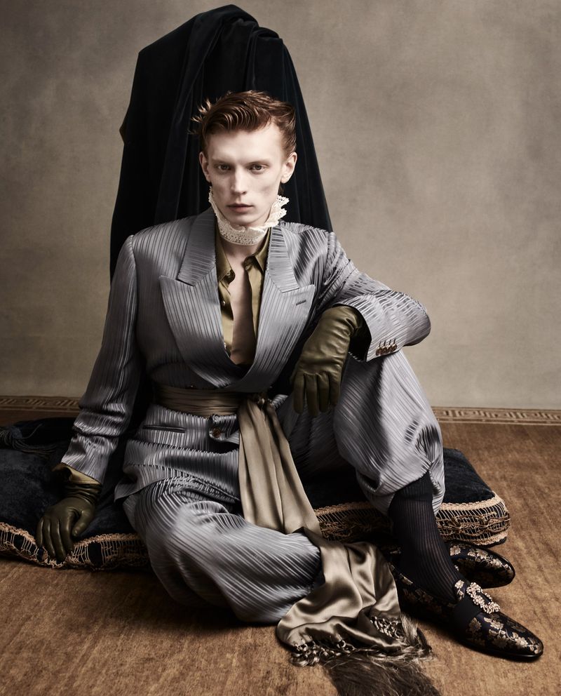 A fair-skinned male model in a grey pinstripe suit with a tan sash sits on a fringed cushion in a studio with soft, neutral lighting.