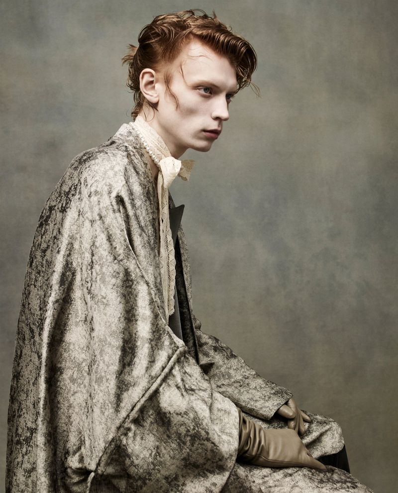 Portrait of a red-haired male model in a mottled grey and white patterned robe and gloves, seated against a mottled grey background.