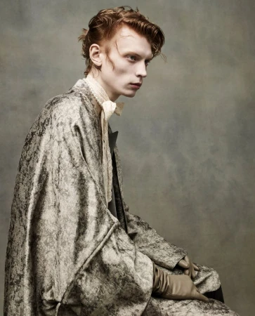 Portrait of a red-haired male model in a mottled grey and white patterned robe and gloves, seated against a mottled grey background.