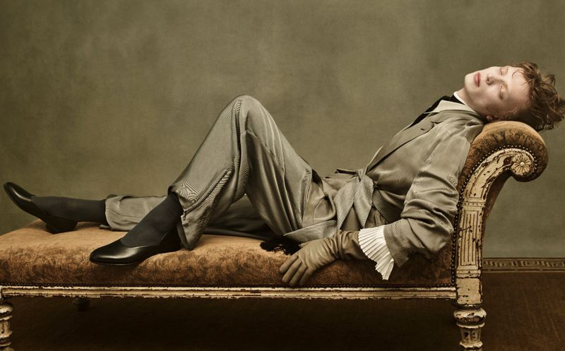 Portrait of a person lying on a chaise lounge in a gray suit and black shoes in front of a muted green background, fashion editorial.