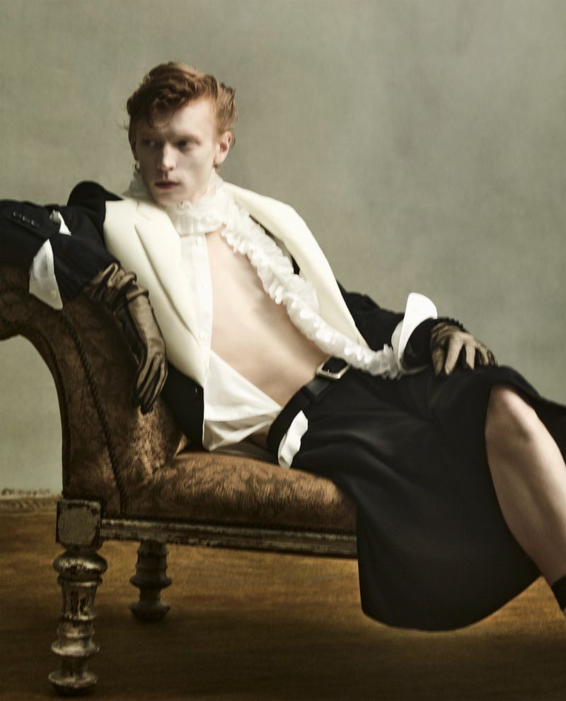 Man with red hair in black jacket, white ruffled shirt, and black skirt reclining on an antique couch indoors in a fashion editorial.