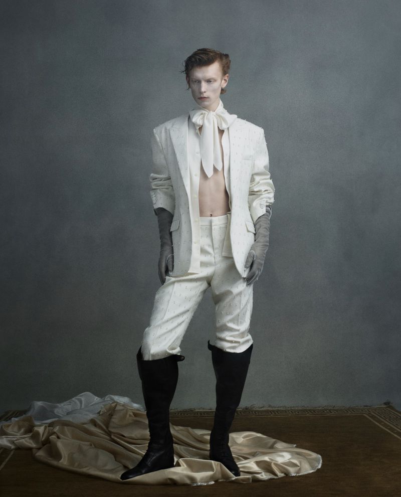 An androgynous person in a white suit, tie, and black boots poses in front of a grey background with draped cream-colored fabric.