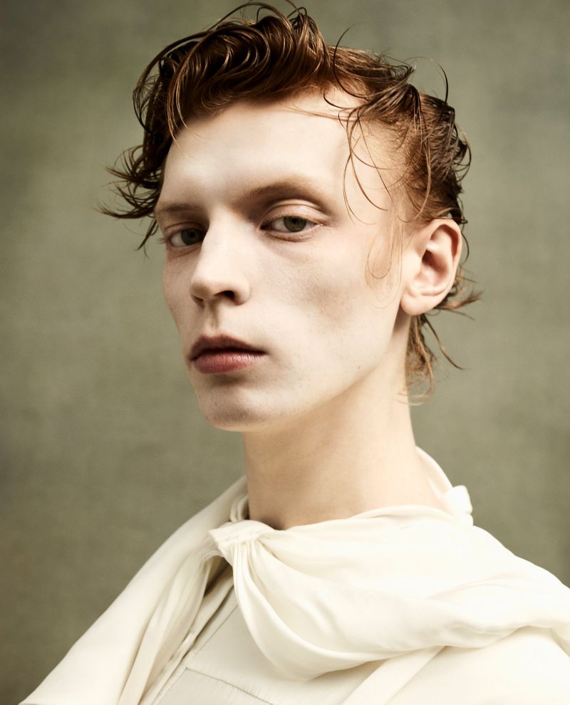 Portrait of a fair-skinned adult male with wet, auburn hair, posing in a cream-colored top against a mottled grey-green backdrop.