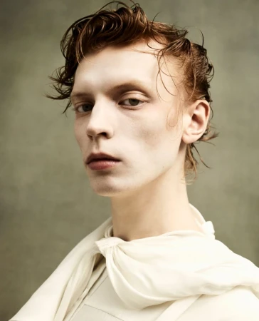 Portrait of a fair-skinned adult male with wet, auburn hair, posing in a cream-colored top against a mottled grey-green backdrop.