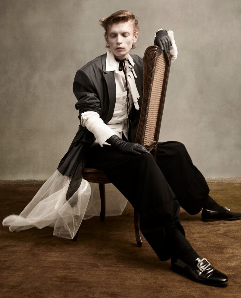 Ethereal fashion portrait of a young adult male in formal wear sitting on a vintage chair against a neutral studio backdrop.
