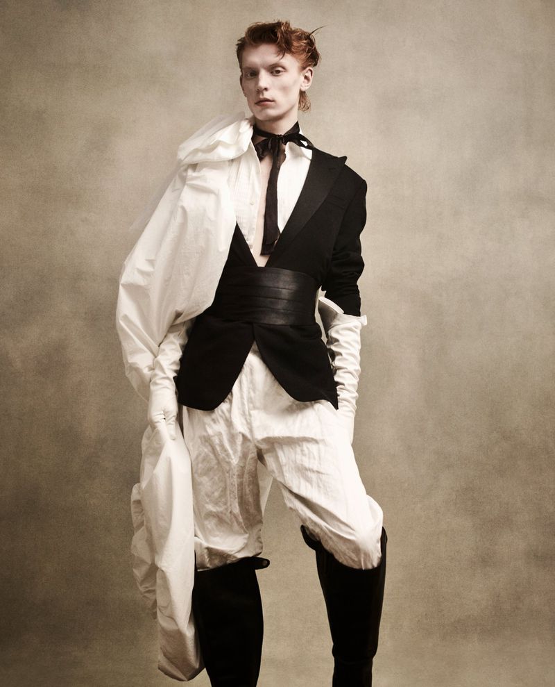 Portrait of a slender person with red hair wearing a black and white outfit with high black boots in a studio setting.