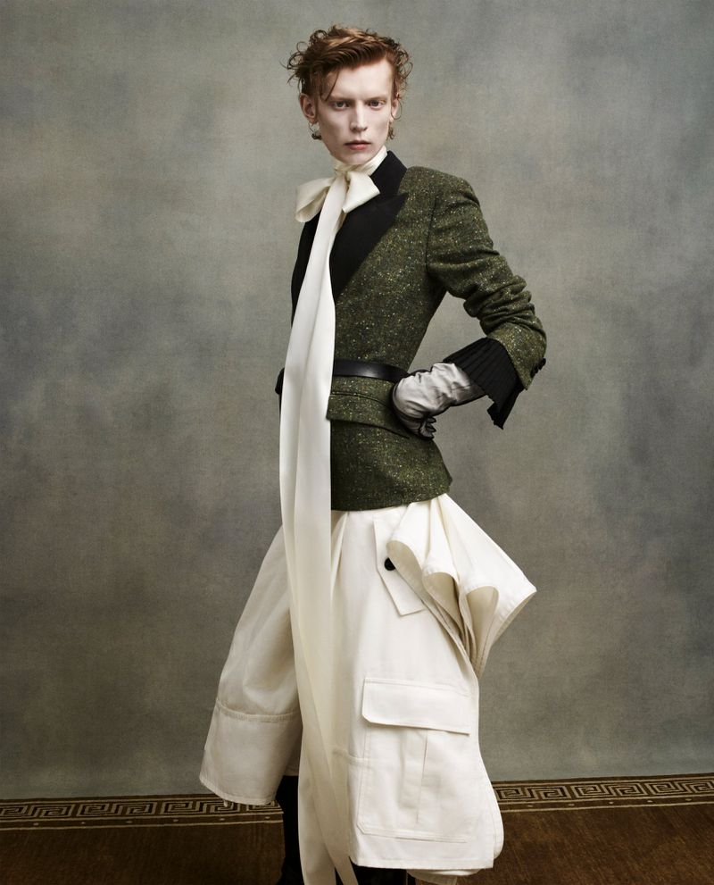 A slender fashion model with curly auburn hair poses in a green jacket, white trousers, and gloves against a gray background in studio lighting.