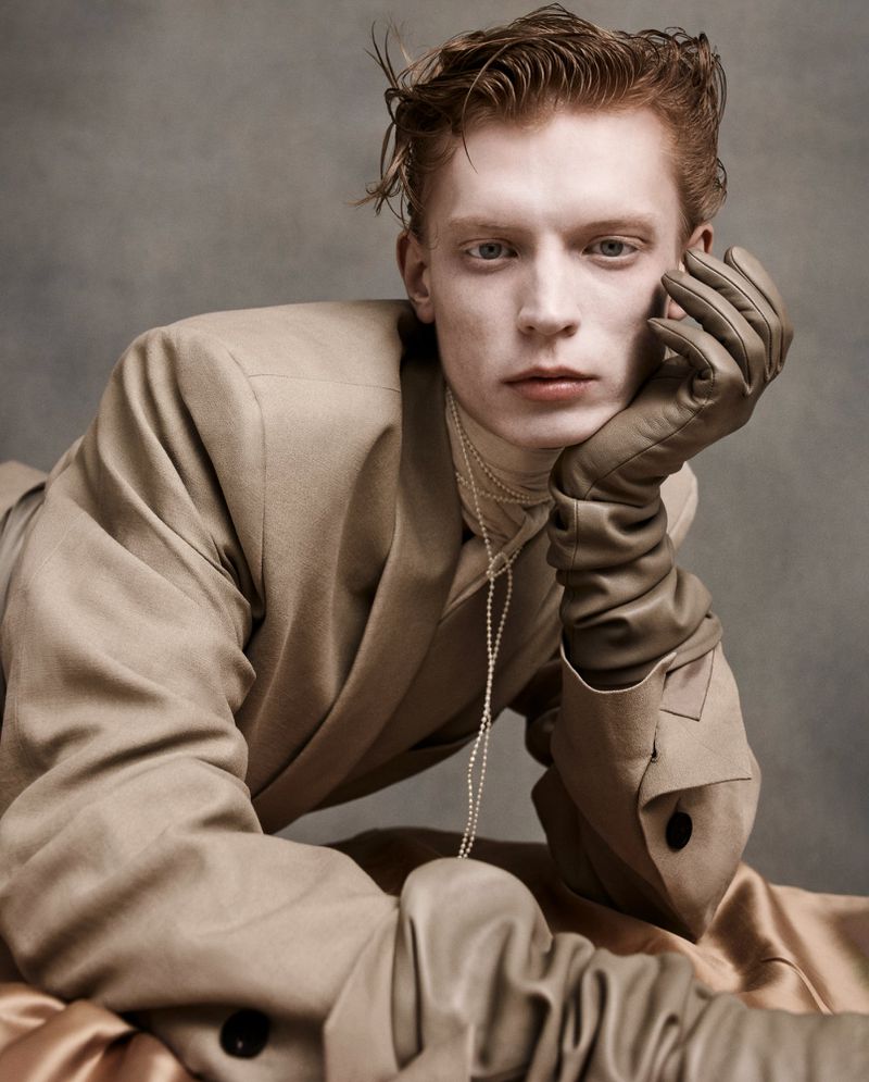 Portrait of a red-haired male model wearing a tan suit and matching gloves, resting his head on his gloved hand against a gray backdrop.