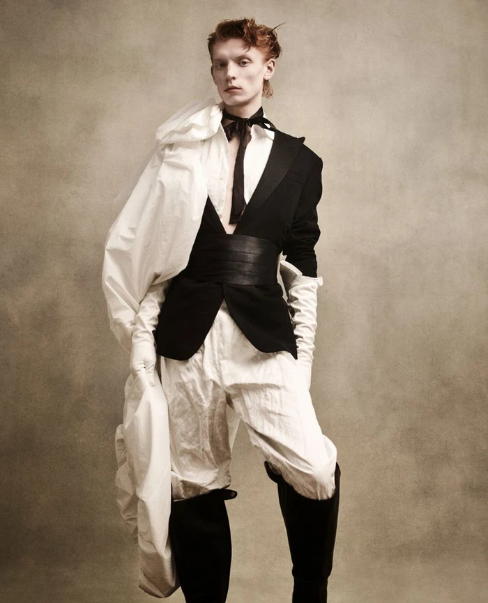 Model Owen Ruppersburg in a black jacket and white trousers with white gloves and black boots against a textured background.