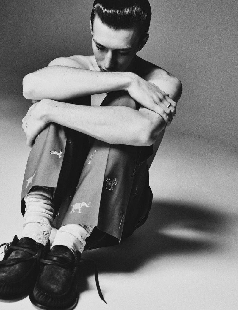 Monochrome studio portrait of a young adult male sitting with arms crossed, wearing patterned pants, socks, and shoes, against a gray backdrop.