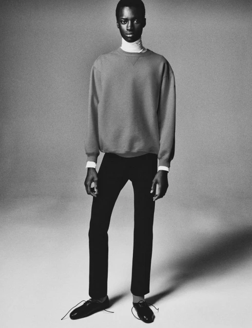 Black and white portrait of an African American male fashion model in a crewneck sweater, pants, and dress shoes posing in a photo studio.