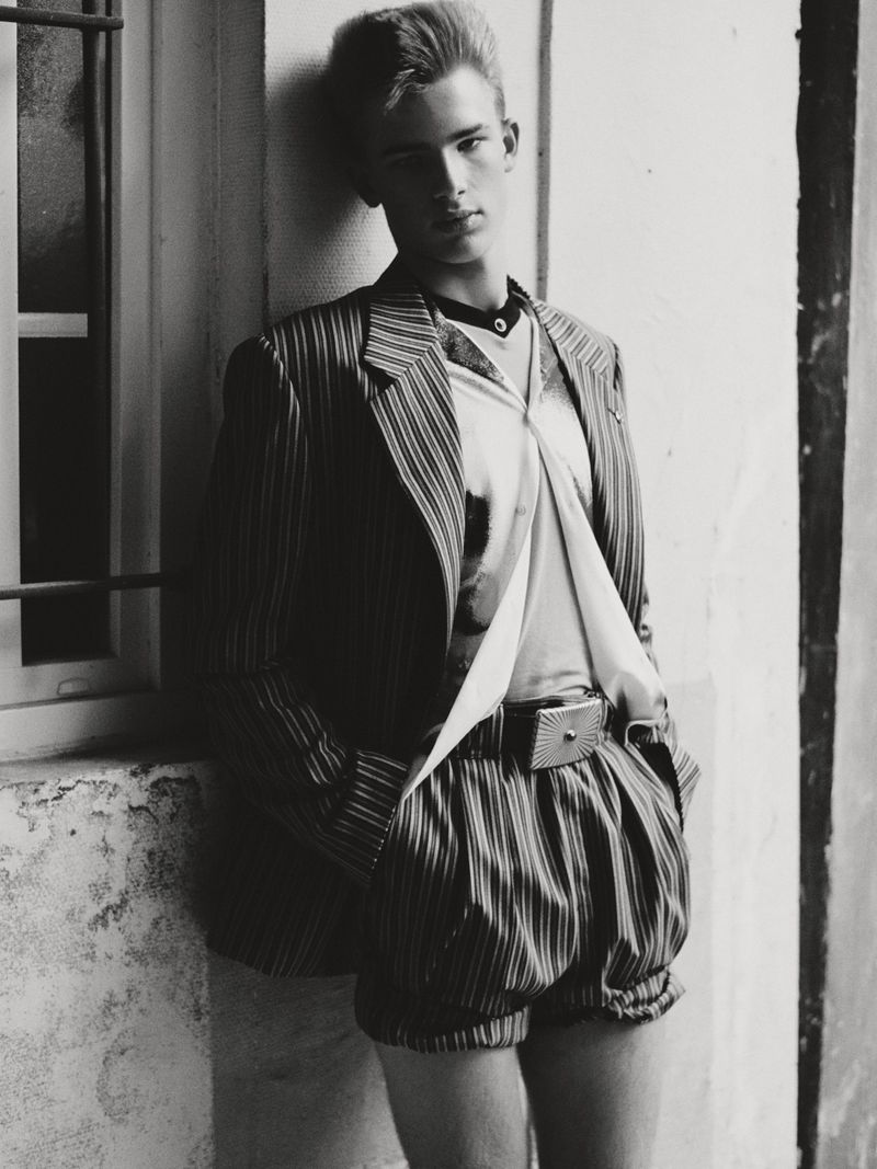 Monochrome portrait of an adult male model in retro pinstripe shorts and jacket, posing against a textured wall and window.