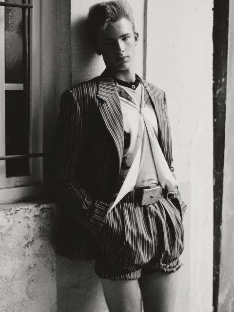 Monochrome portrait of an adult male model in retro pinstripe shorts and jacket, posing against a textured wall and window.
