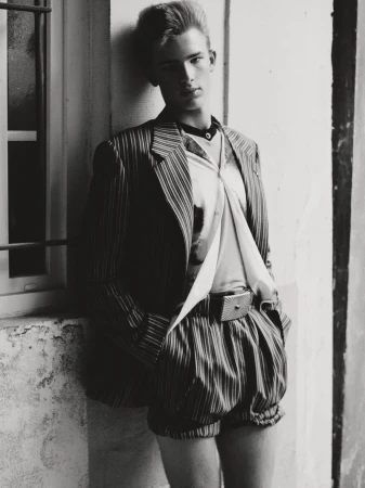 Monochrome portrait of an adult male model in retro pinstripe shorts and jacket, posing against a textured wall and window.