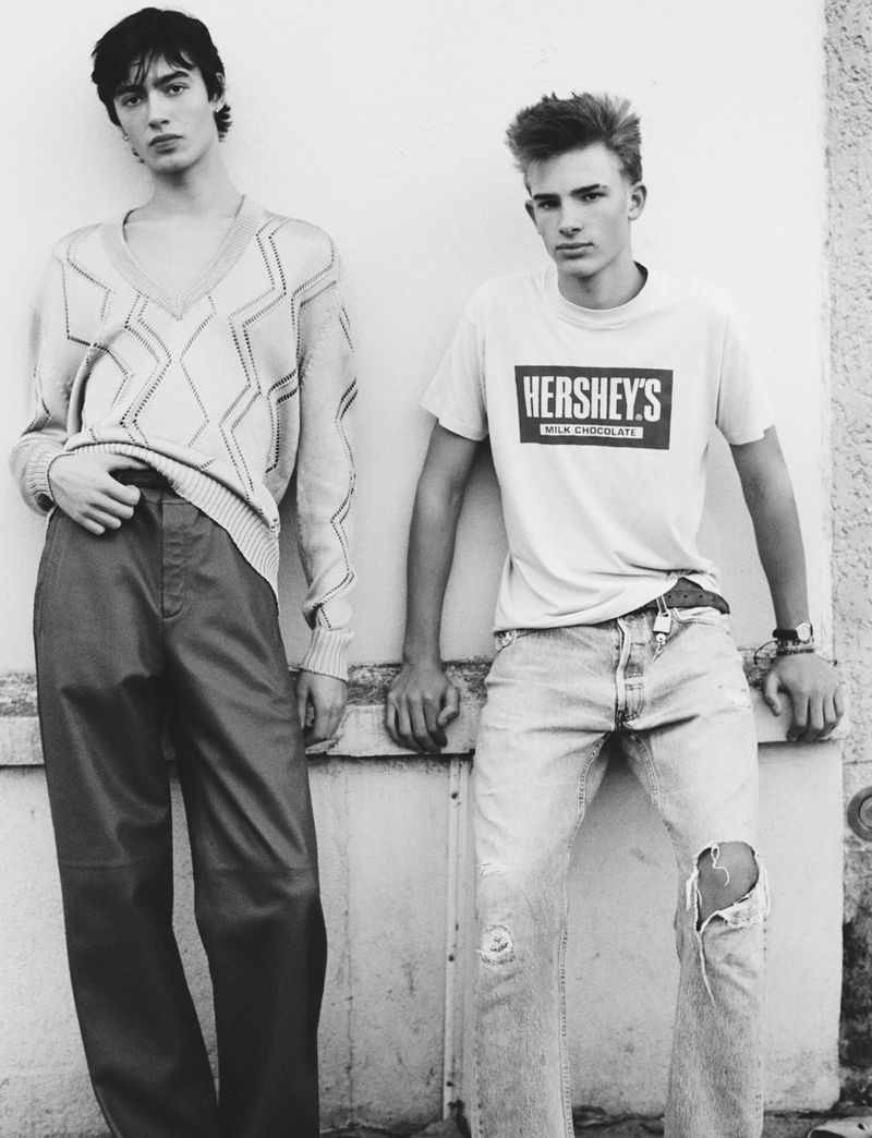 Two young men pose indoors, one in a patterned sweater and pants, the other in a t-shirt with a Hershey's logo and ripped jeans.