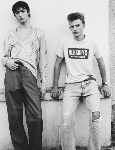 Two young men pose indoors, one in a patterned sweater and pants, the other in a t-shirt with a Hershey's logo and ripped jeans.