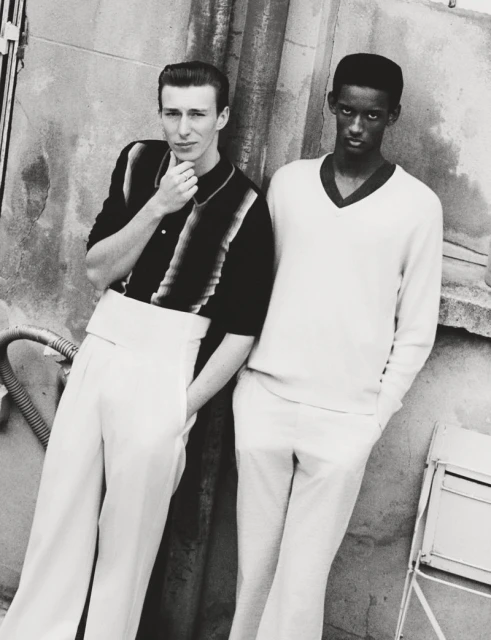 Monochromatic fashion shot of two young men in 1950s style attire, standing against a weathered wall with pipes and a small metal table.