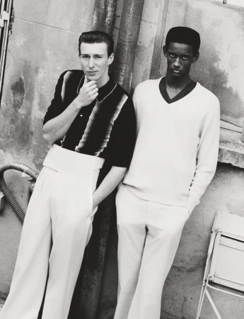 Monochromatic fashion shot of two young men in 1950s style attire, standing against a weathered wall with pipes and a small metal table.