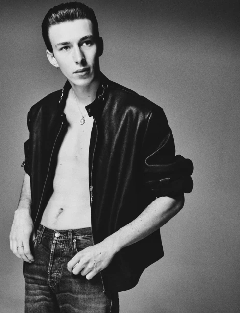 Monochrome portrait of a young adult man with slicked-back hair, wearing a leather jacket and jeans against a plain background.