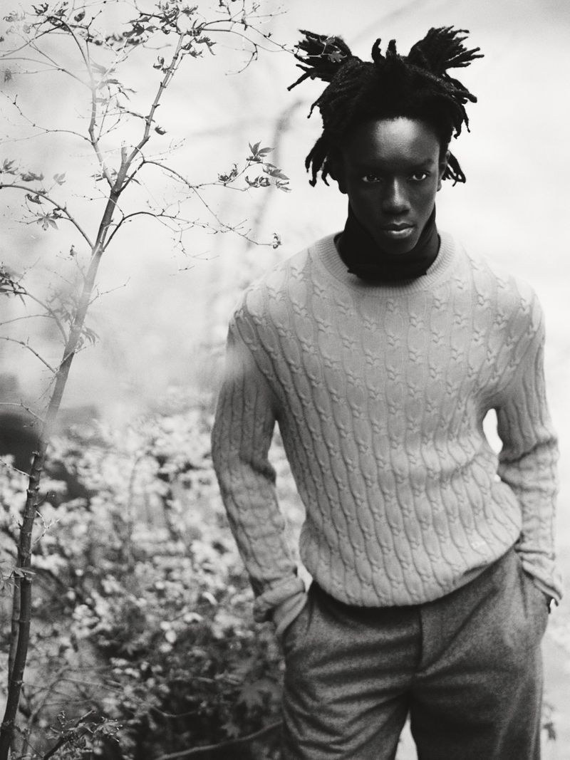 Black man wearing textured knit sweater and dark trousers standing outdoors near bare branches.