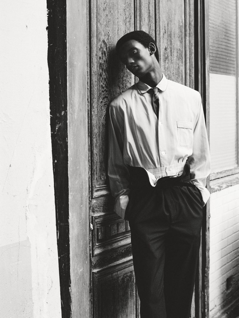 Monochrome portrait of a Black man leaning against an old wooden door wearing a button-down shirt, tie, and slacks in an outdoor setting.
