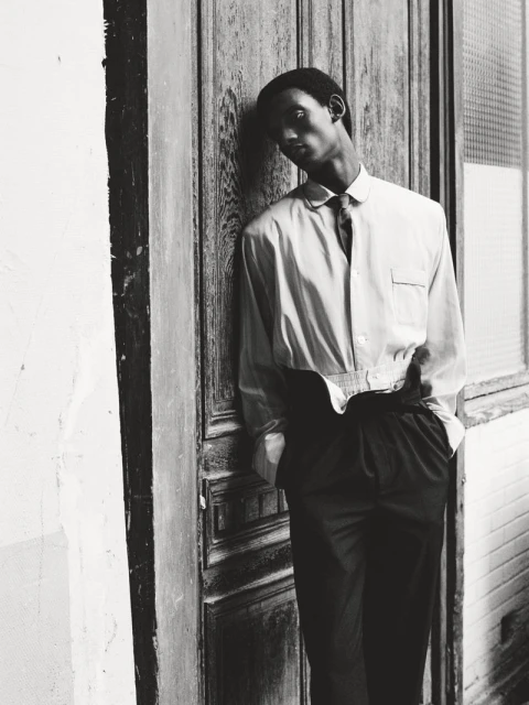 Monochrome portrait of a Black man leaning against an old wooden door wearing a button-down shirt, tie, and slacks in an outdoor setting.