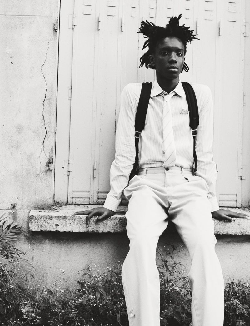 Black and white portrait of a young man with dreadlocks sitting outside wearing a shirt, tie, and suspenders, with plants visible in the foreground.