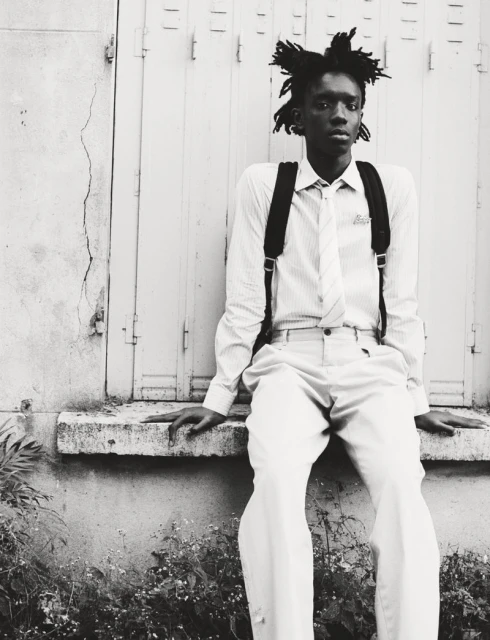 Black and white portrait of a young man with dreadlocks sitting outside wearing a shirt, tie, and suspenders, with plants visible in the foreground.