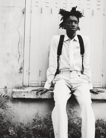 Black and white portrait of a young man with dreadlocks sitting outside wearing a shirt, tie, and suspenders, with plants visible in the foreground.