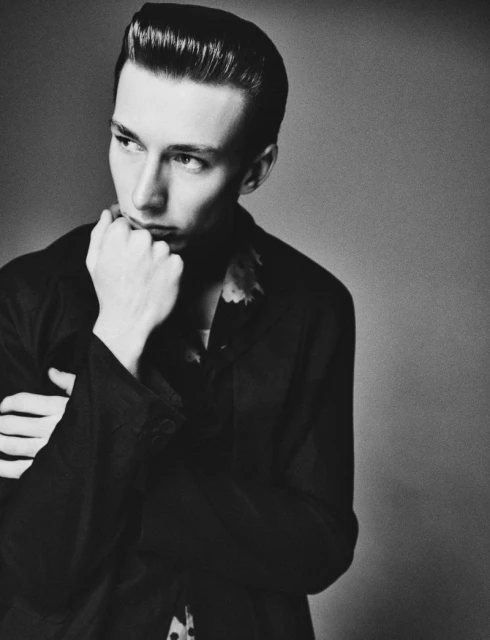 Black and white portrait of an adult male with slicked-back hair, wearing a dark jacket, hand on his chin, looking off to the side.