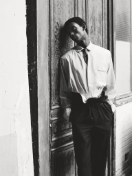Black and white editorial fashion portrait of a male model leaning against a wooden door wearing trousers and a collared shirt