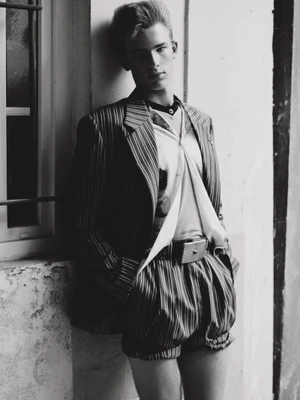 Young man in striped blazer and shorts leaning against an exterior wall for a fashion editorial portrait