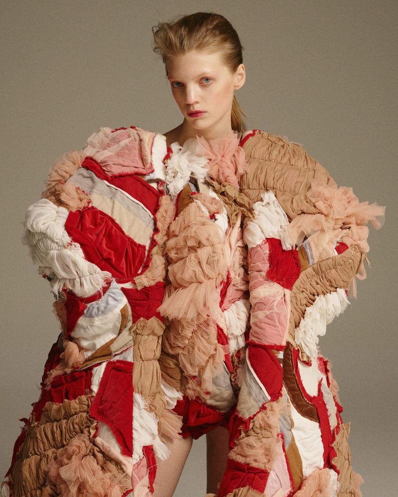 Fashion model in a photo studio. She is wearing an avant-garde patchwork dress made of red, beige and white textured fabrics. 