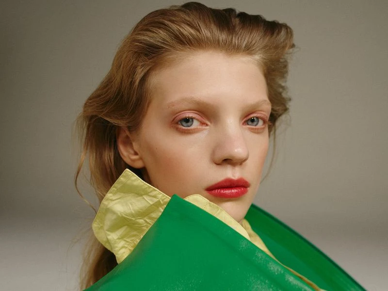 Close-up portrait of a fair-skinned woman with blonde hair, red lipstick, wearing a lime-green and yellow top against a gray backdrop.