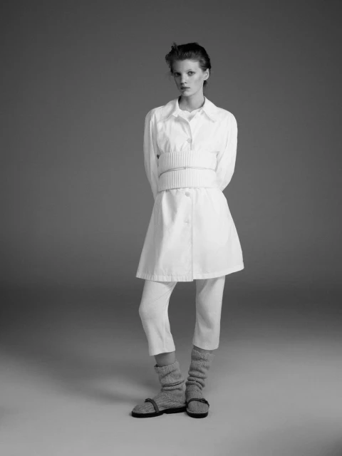 Monochrome portrait of a young woman in an all-white ensemble and layered sweater, set against a neutral gray studio backdrop.