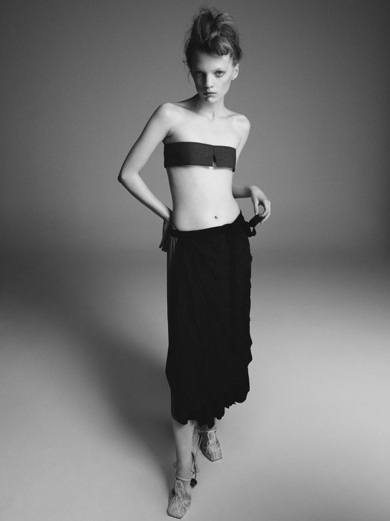 Monochrome fashion shot of a slender woman in a strapless top and draped skirt, striking a pose in a neutral studio setting.