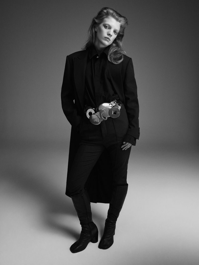 A woman in a black outfit poses in a studio wearing a belt with metallic objects. The black and white shot has a neutral grey background.