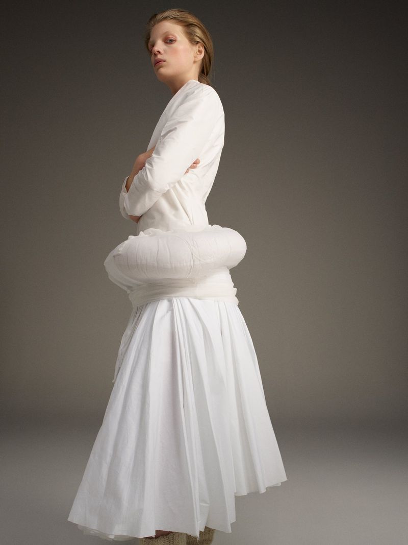 Blonde woman in white dress poses in studio with neutral background. Fashion shot shows full-length view of designer outfit.