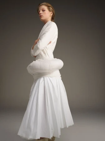 Blonde woman in white dress poses in studio with neutral background. Fashion shot shows full-length view of designer outfit.