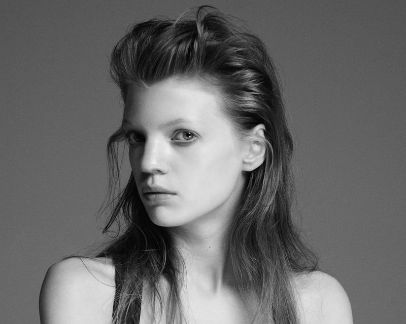 Striking black and white portrait of a young woman with styled hair, gazing directly at the camera against a neutral backdrop.