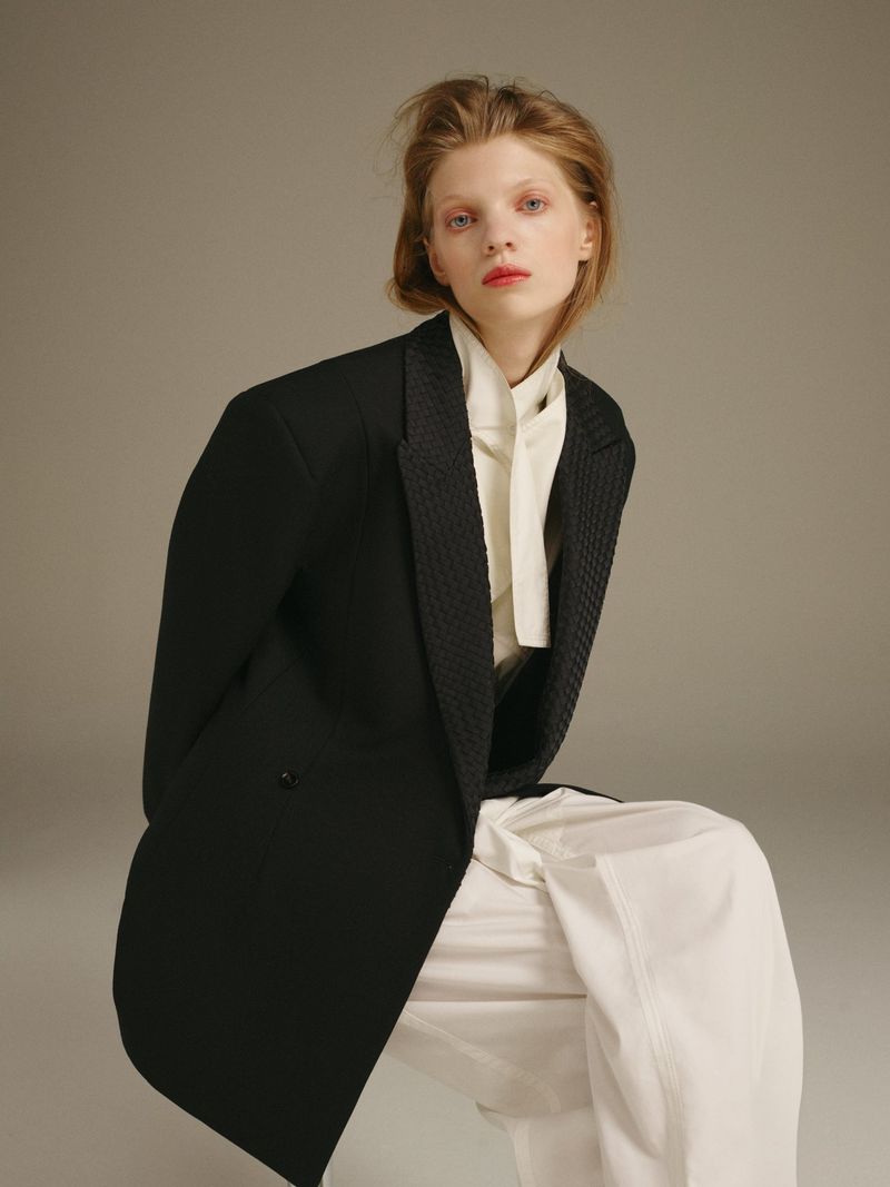 A blond-haired woman in a black blazer and white pants poses in a fashion shot against a neutral grey background.