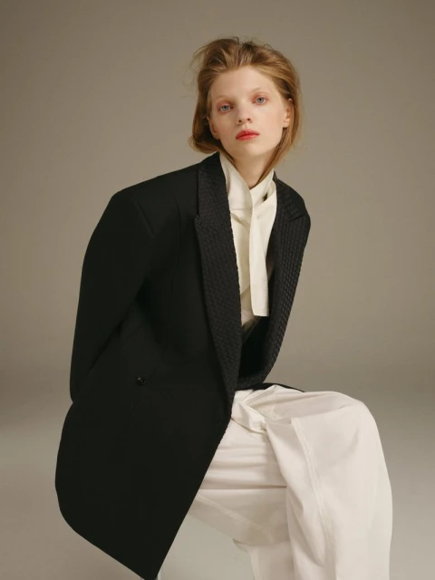 A blond-haired woman in a black blazer and white pants poses in a fashion shot against a neutral grey background.